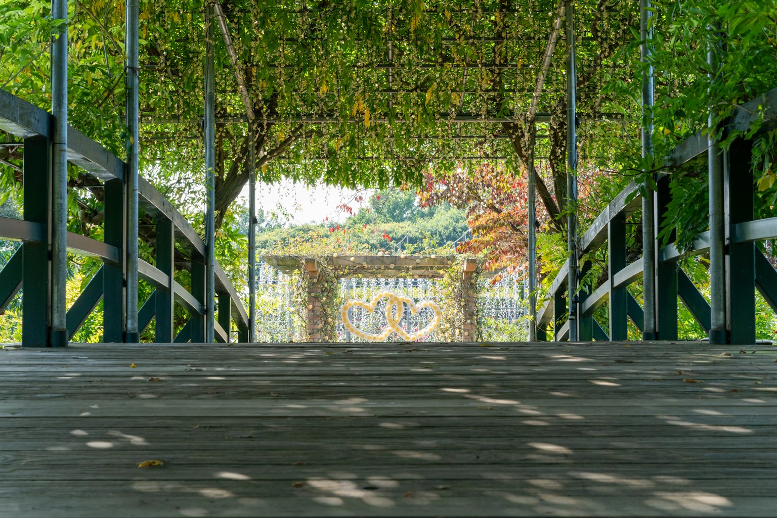 Blick auf eine Holzbrücke, die durch einen grünen Bogen führt, mit Sonnenlicht, das hindurchscheint