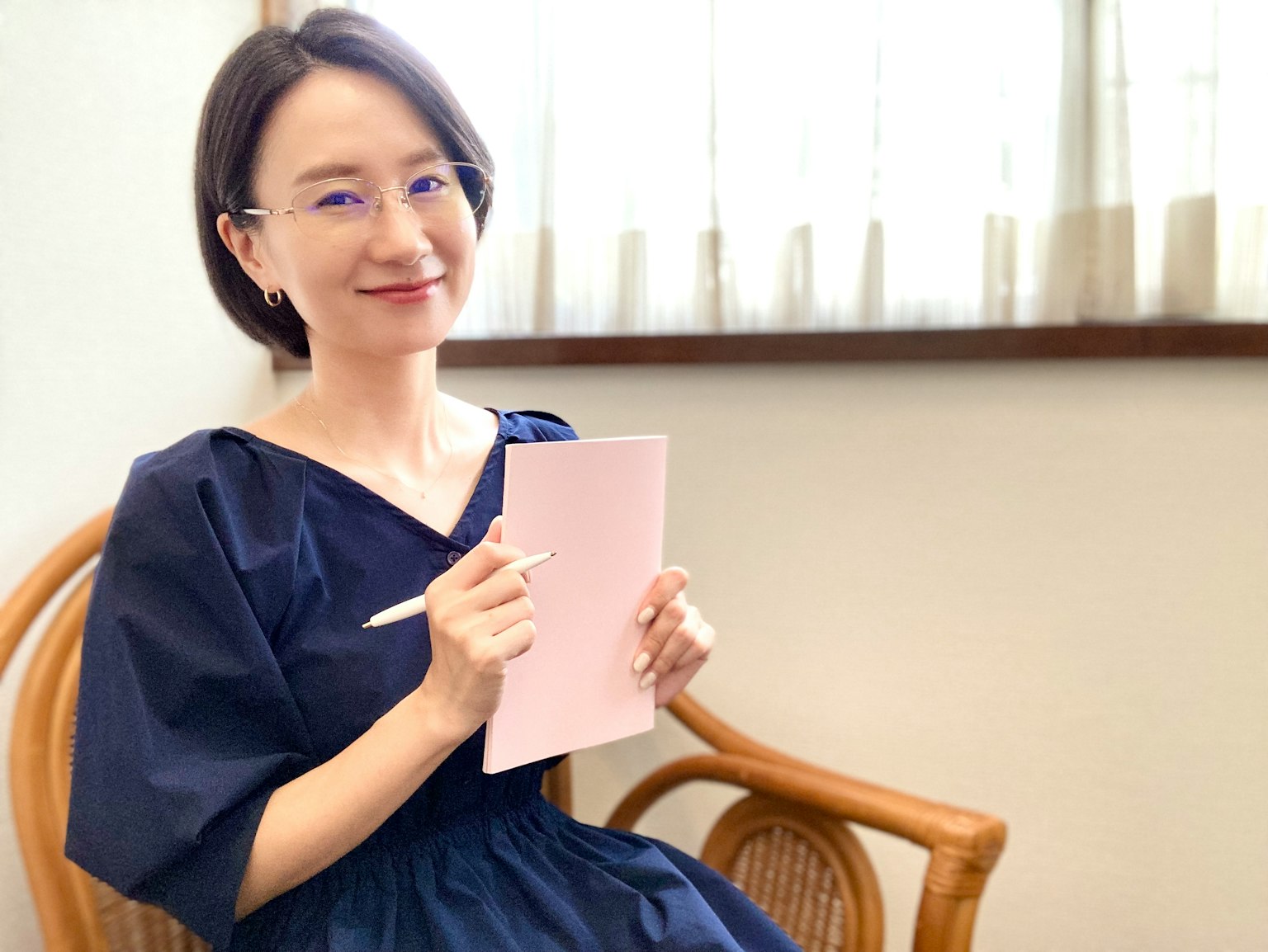 Una mujer con un vestido azul sonriendo mientras sostiene un cuaderno rosa y un bolígrafo