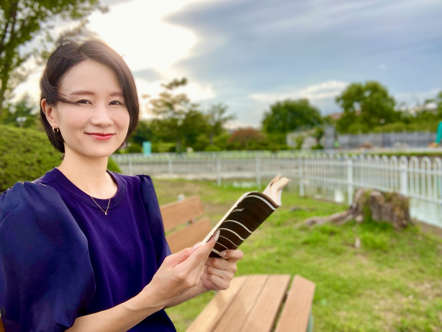 Wanita memegang buku di taman dengan langit cerah dan latar belakang hijau