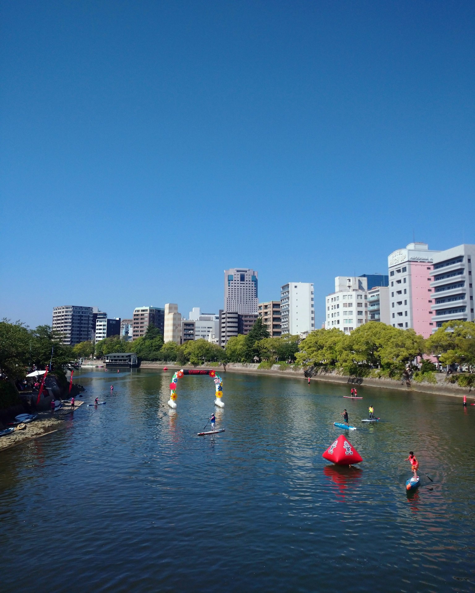 在蓝天的映衬下，人们在河上享受划皮艇和立式划水，背景是城市风景
