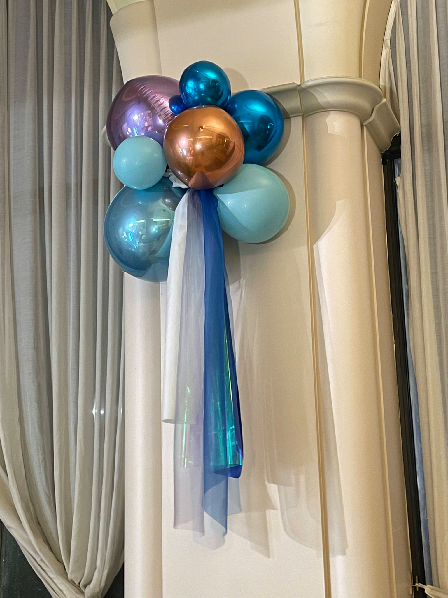 Un arreglo decorativo de globos azules, morados y dorados con cintas atadas a una columna