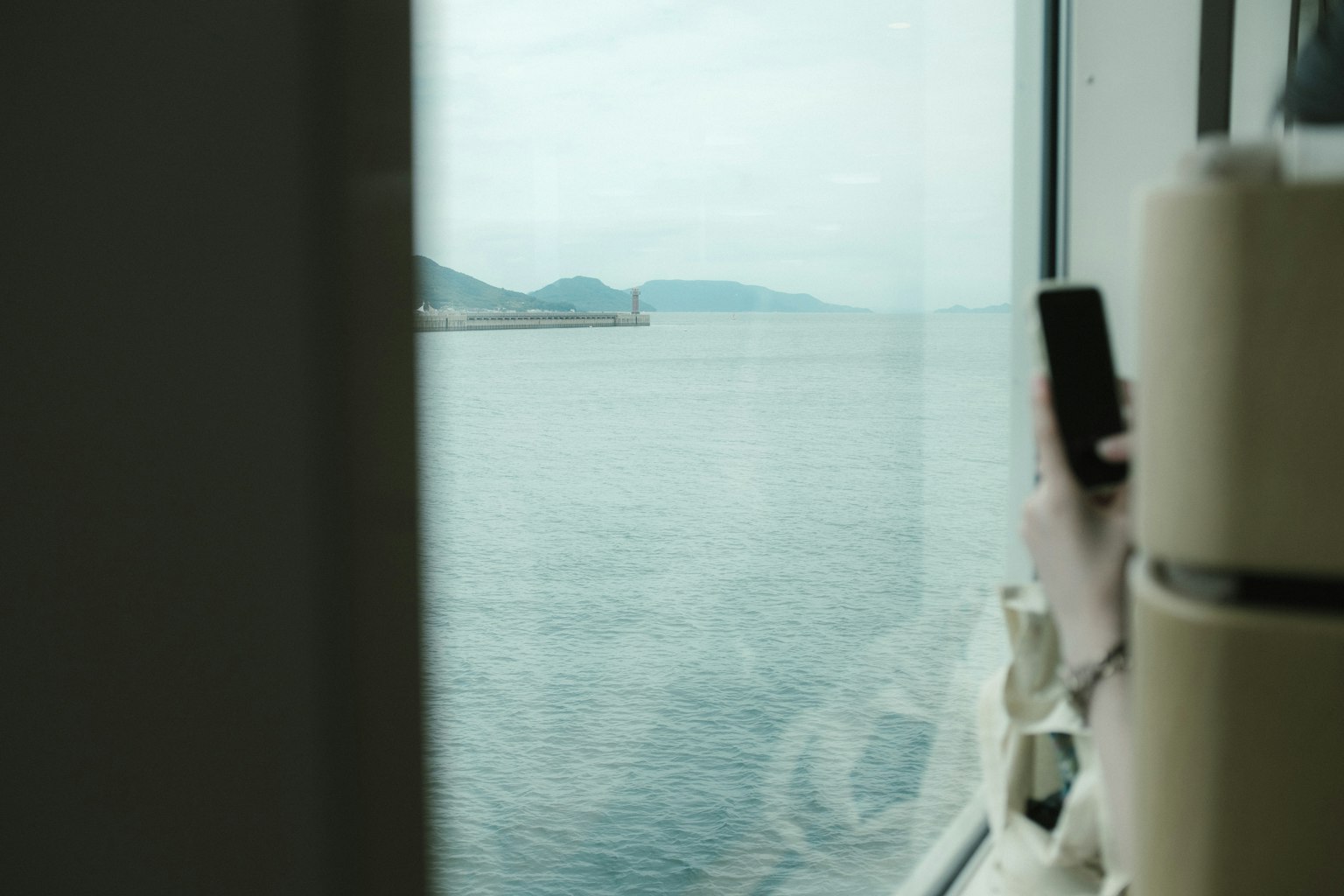 Vista del mare e delle montagne attraverso una finestra con una persona che tiene uno smartphone