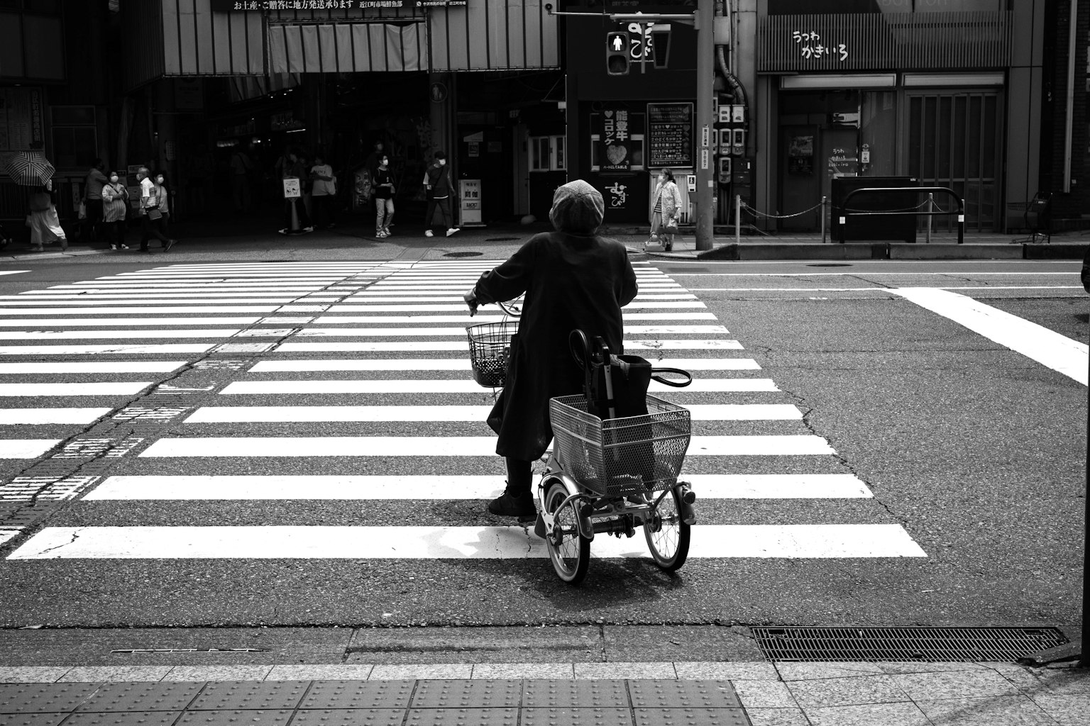 Schwarzweißfoto einer älteren Person, die mit einem Einkaufswagen einen Zebrastreifen überquert