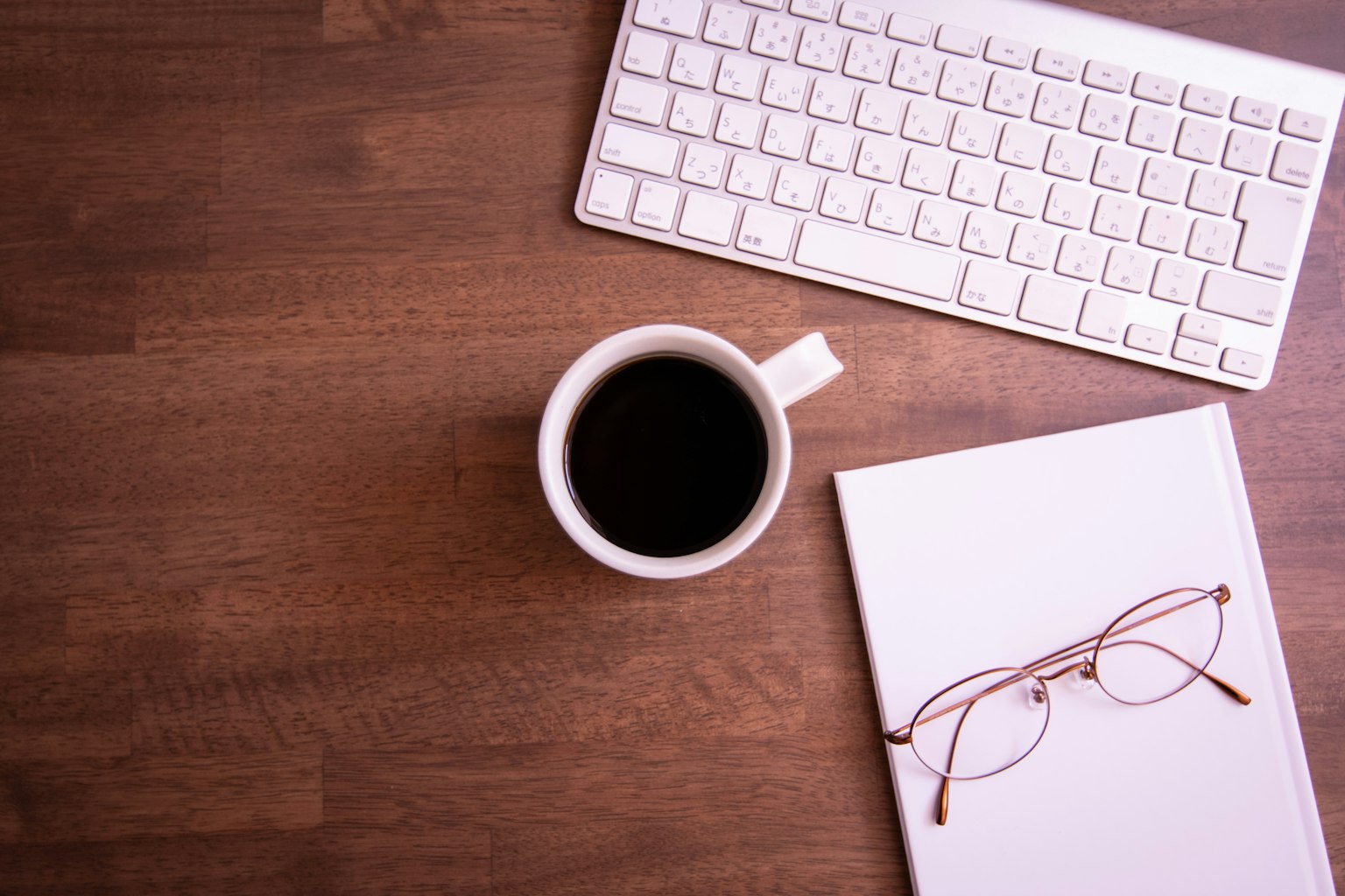 나무 테이블 위에 놓인 흰색 키보드와 커피 컵 노트북과 안경이 옆에 있음