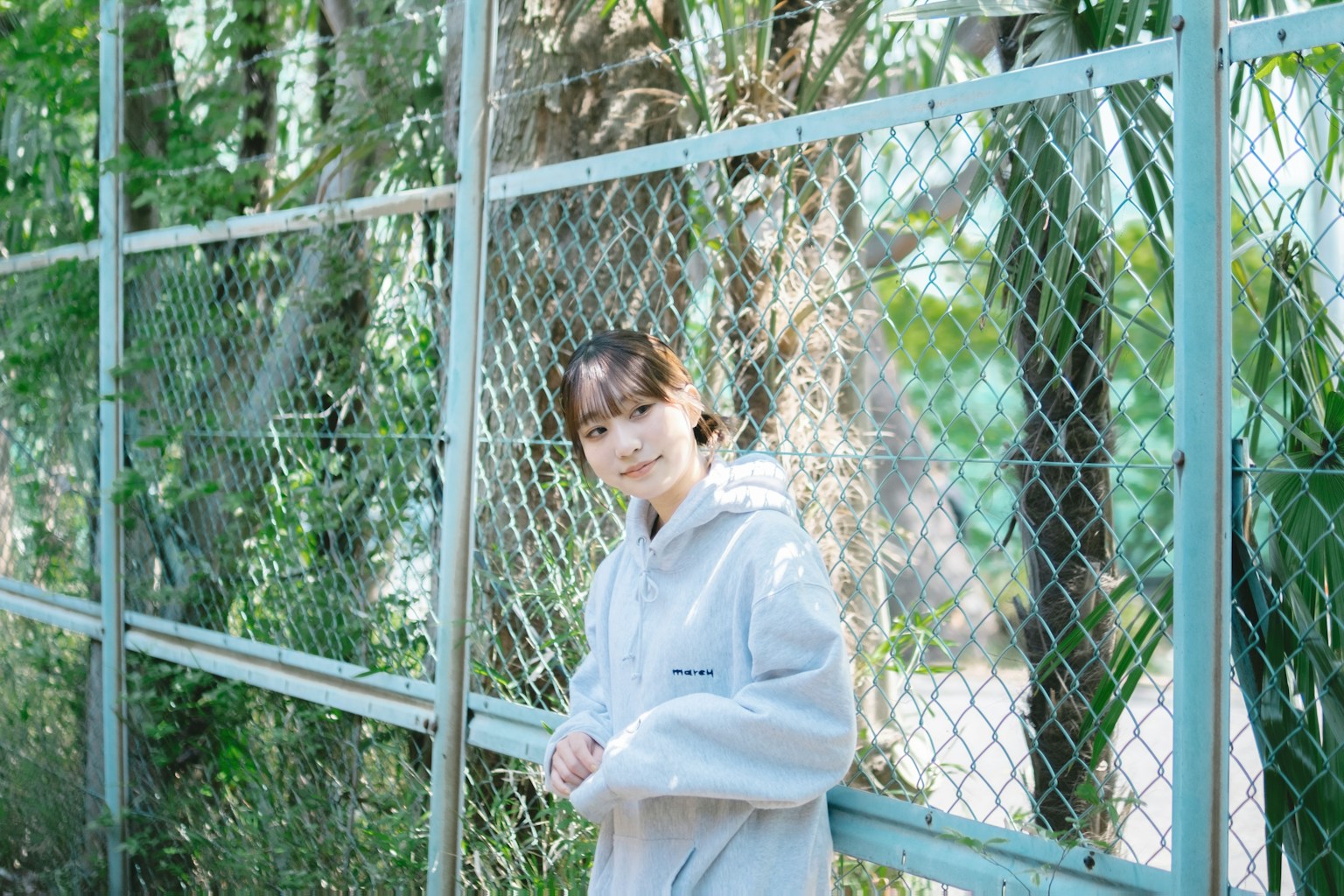 A woman smiling in front of a fence She wears a gray hoodie with green plants in the background