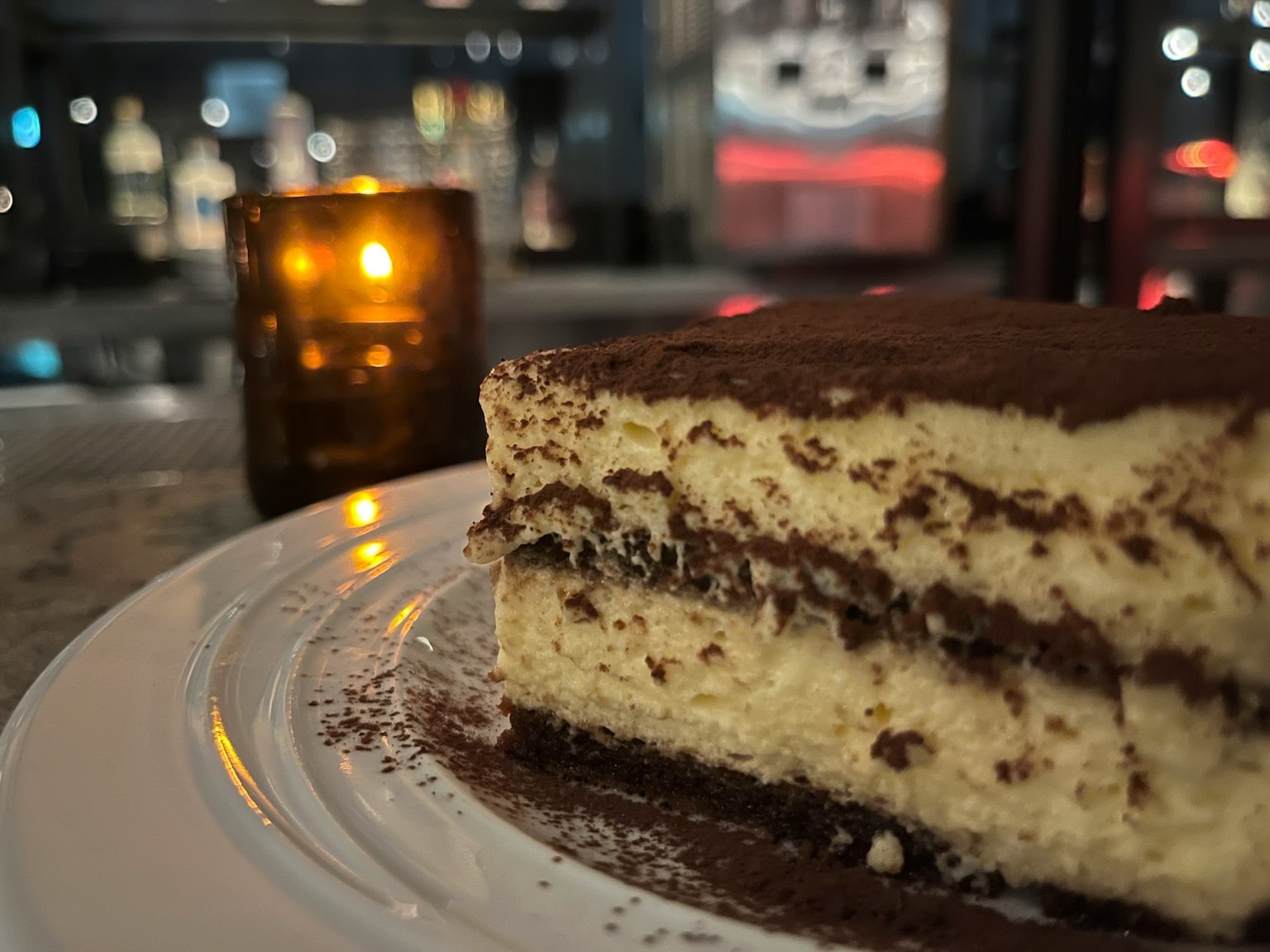 Side view of tiramisu cake on a plate with a candle glowing in the background