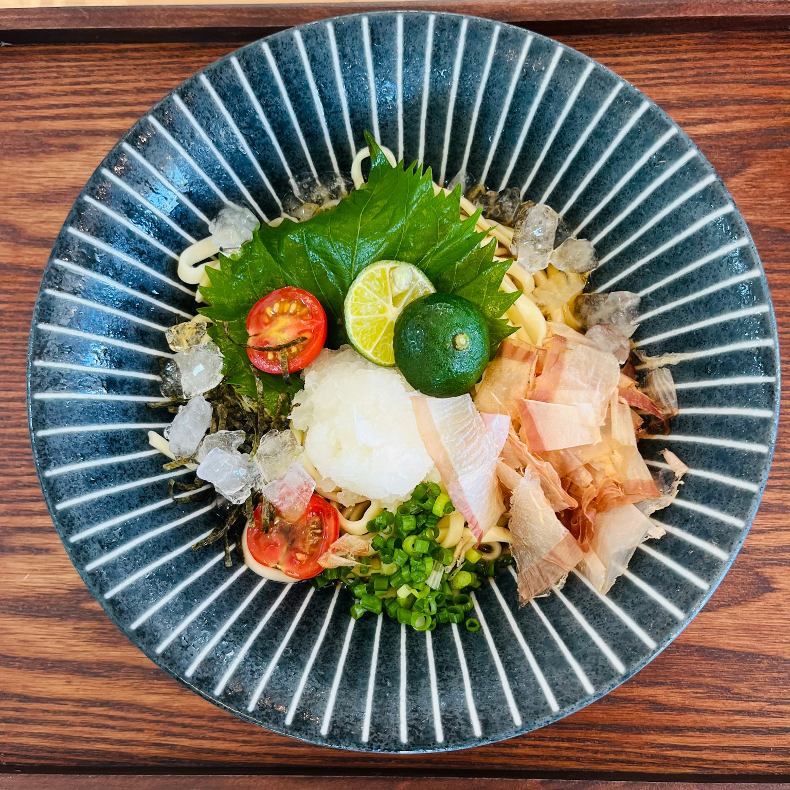 아름답게 담아낸 일본식 냉면, 파란 줄무늬 그릇에 토마토, 쪽파, 무채, 시소잎, 감귤이 곁들여져 있음