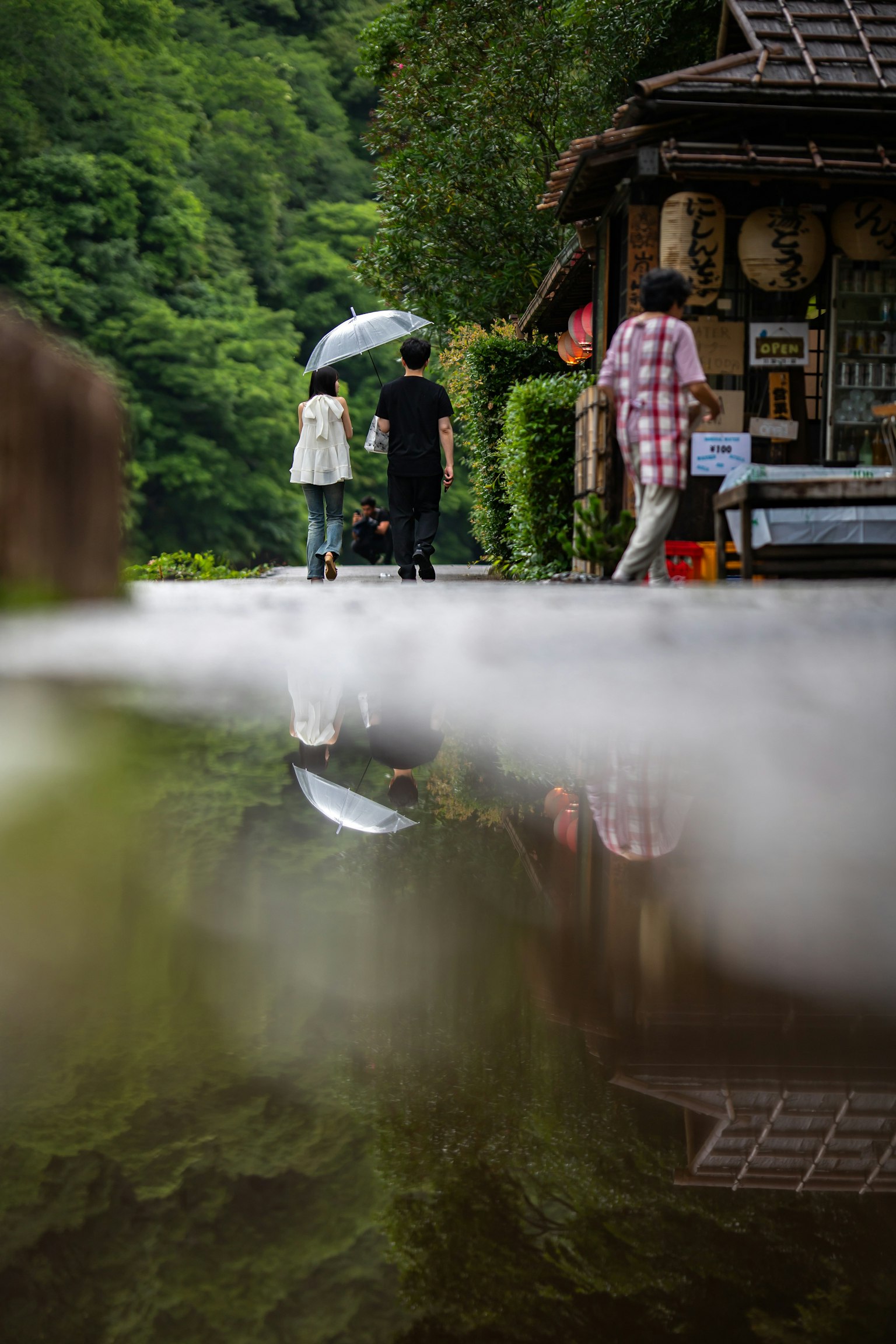 Personas caminando con un paraguas en un paisaje verde reflejado en un charco