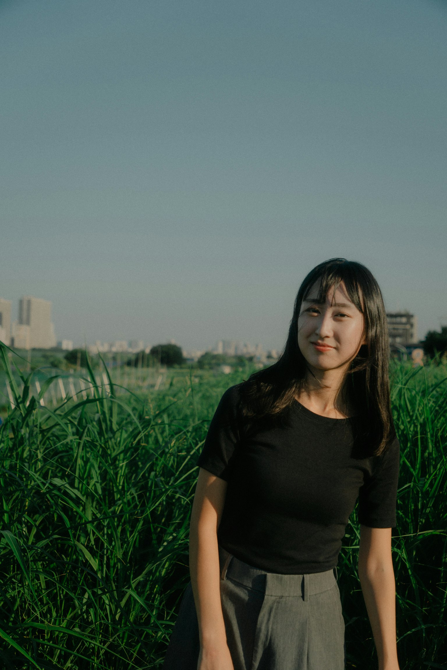 草地上站着的女性肖像，穿着黑色T恤和灰色裤子，背景是城市风景