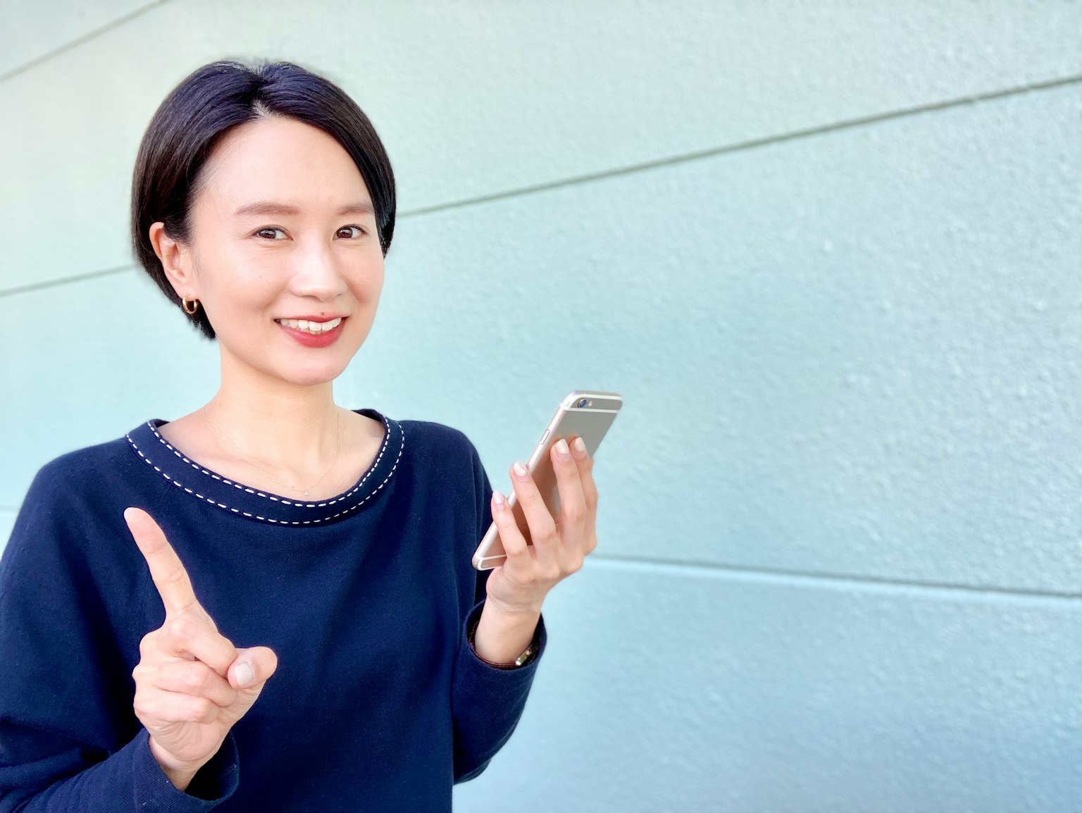 Una donna in piedi davanti a un muro blu che tiene uno smartphone e sorride con un dito alzato