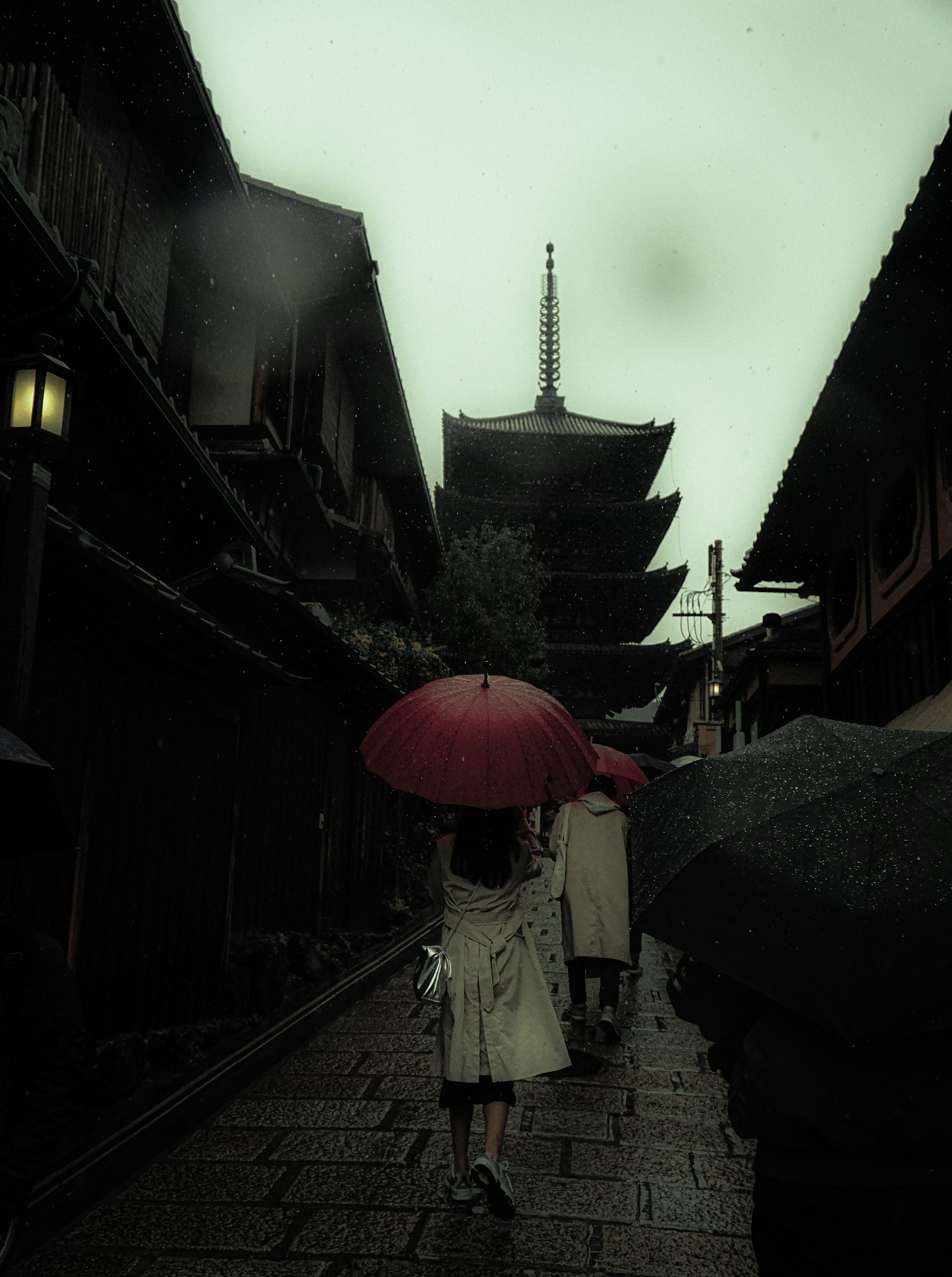 A woman holding a red umbrella walks through an old street with a pagoda in the background on a rainy day