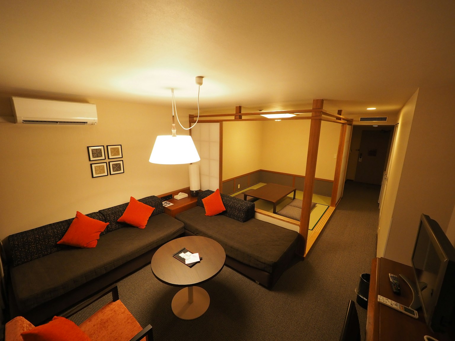 Gemütliches Wohnzimmer mit einem Ecksofa mit orangefarbenen Kissen einem runden Couchtisch und einem Eingang zu einem Tatami-Zimmer