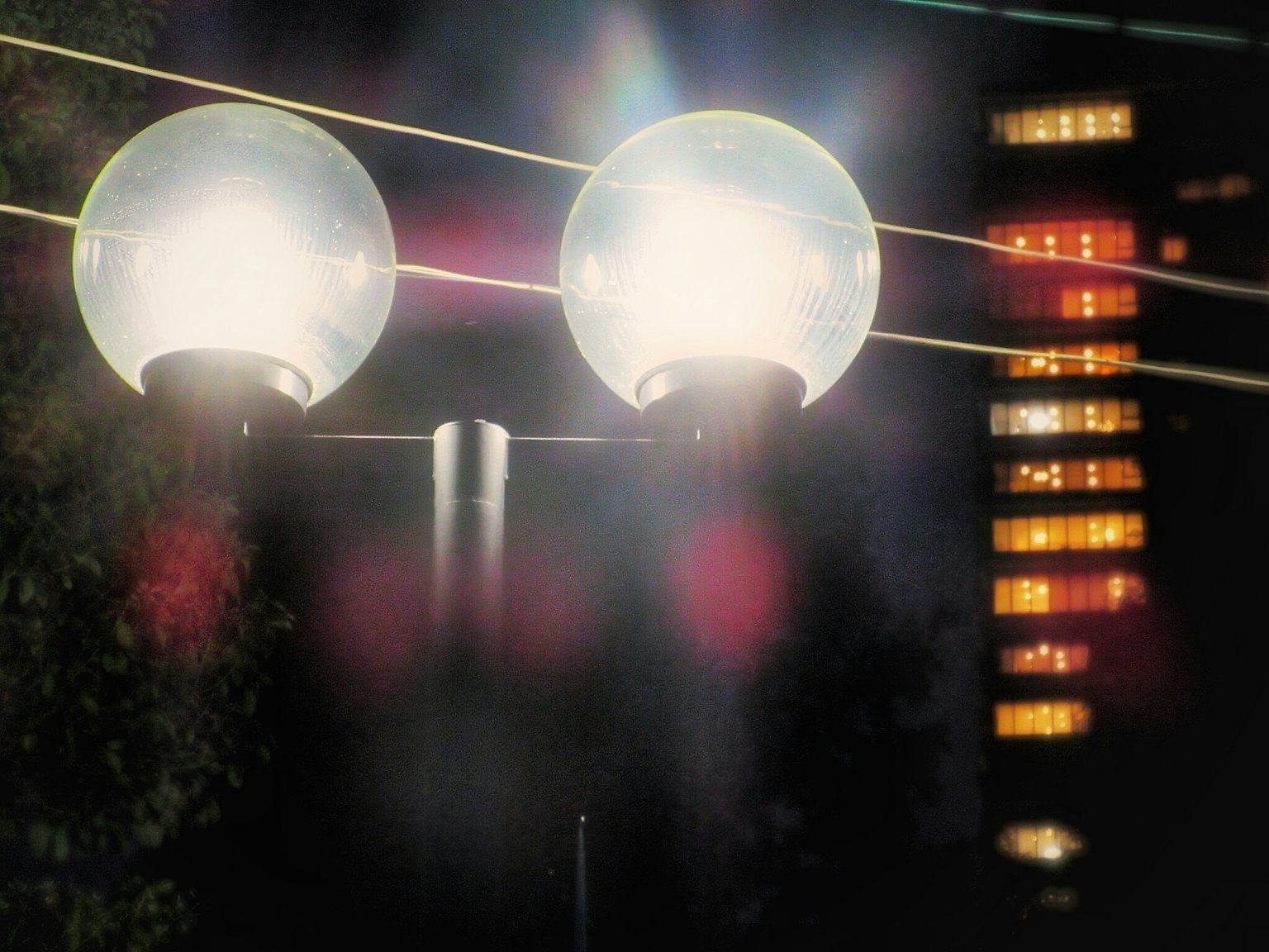 Close-up lampu jalan di malam hari dengan dua lampu bulat terang dan gedung tinggi yang buram di latar belakang