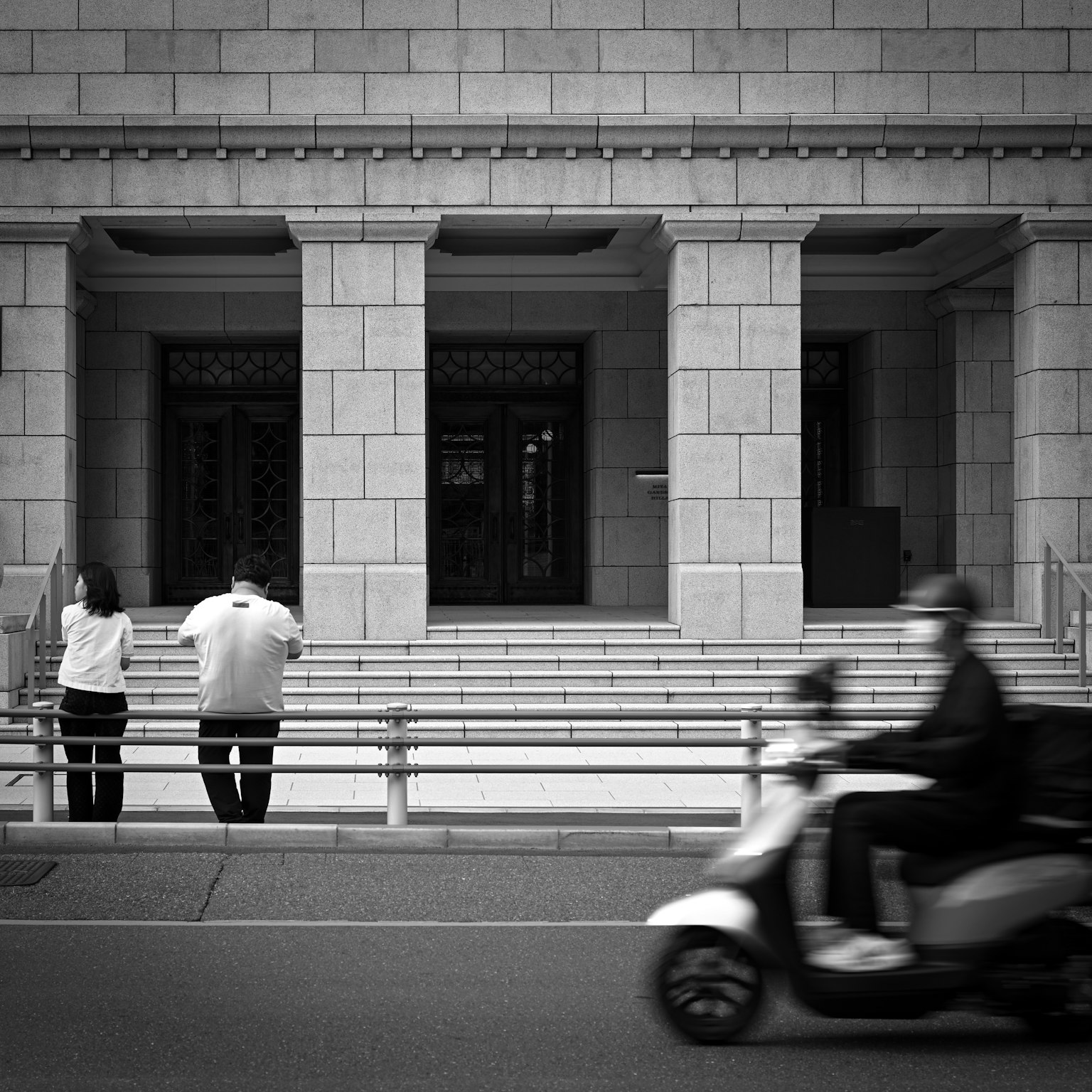 Scena di strada in bianco e nero con persone in piedi e uno scooter che passa