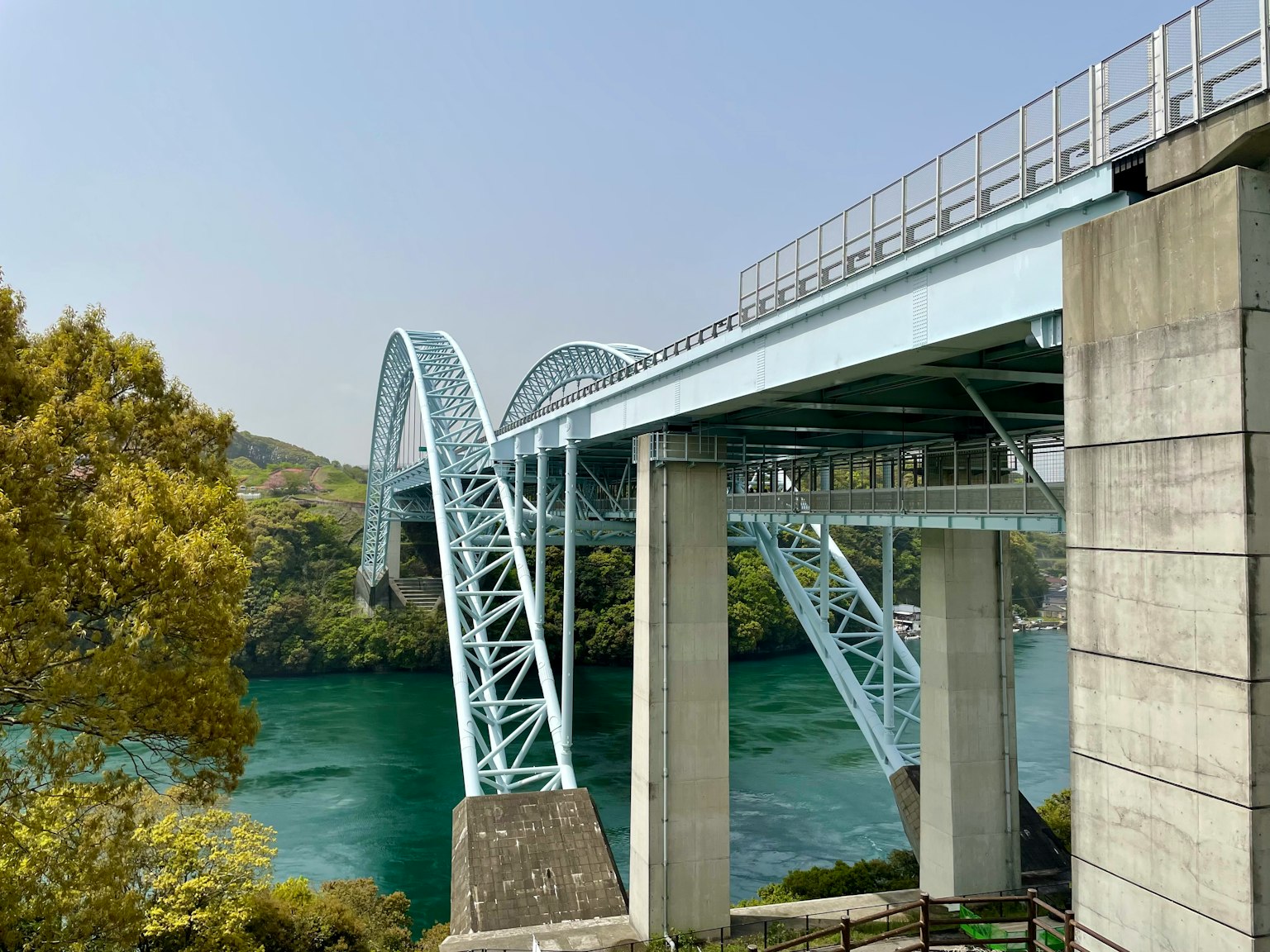 A blue bridge spanning over a river with concrete supports beneath surrounded by green trees
