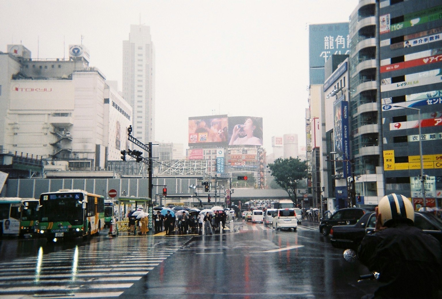 雨中的城市景觀，公車和汽車在交叉口穿行，背景有大型廣告