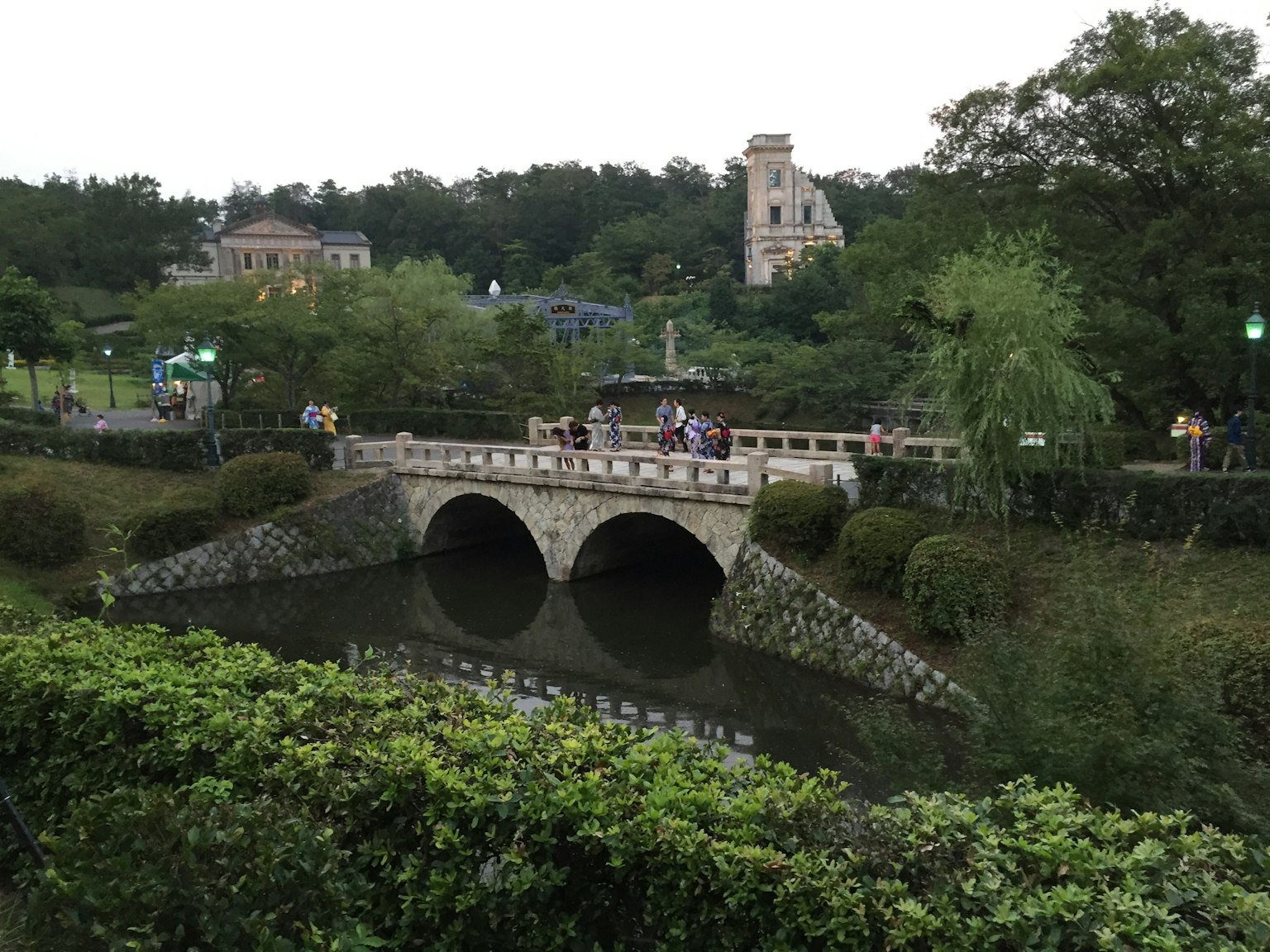 Escena de parque con un puente de piedra sobre un estanque y personas