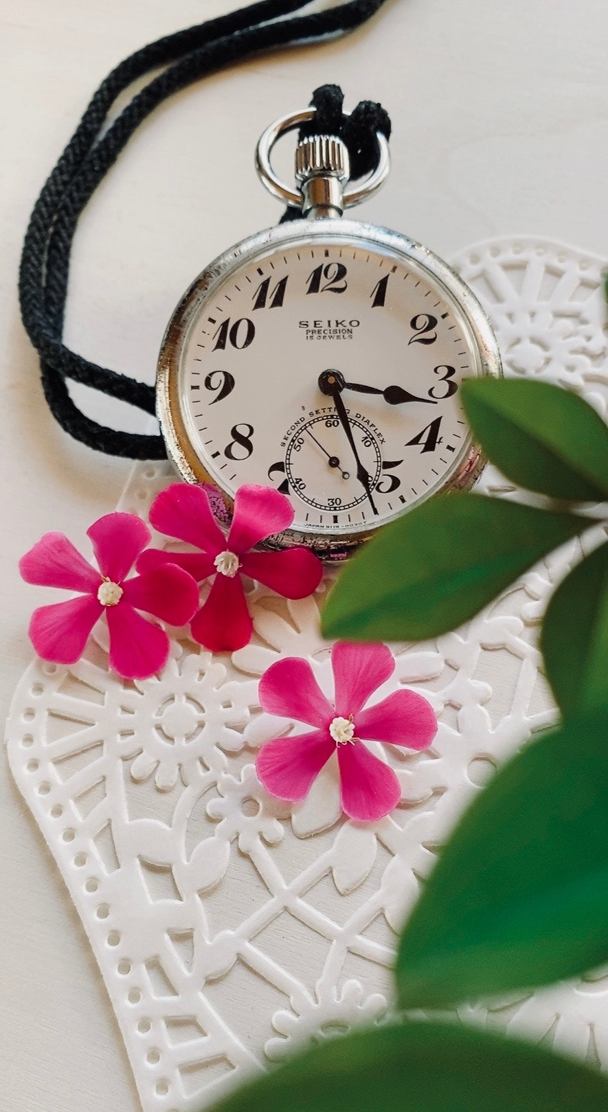 A silver pocket watch beside pink flowers on a decorative lace background