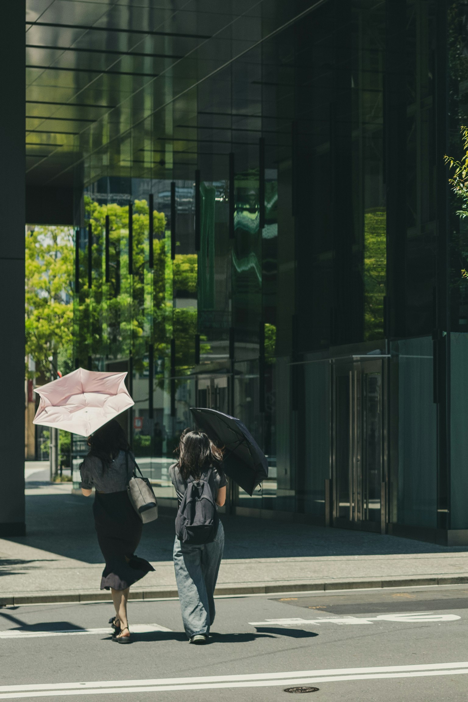 Dua wanita berjalan dengan payung di depan gedung modern dengan pohon hijau di latar belakang