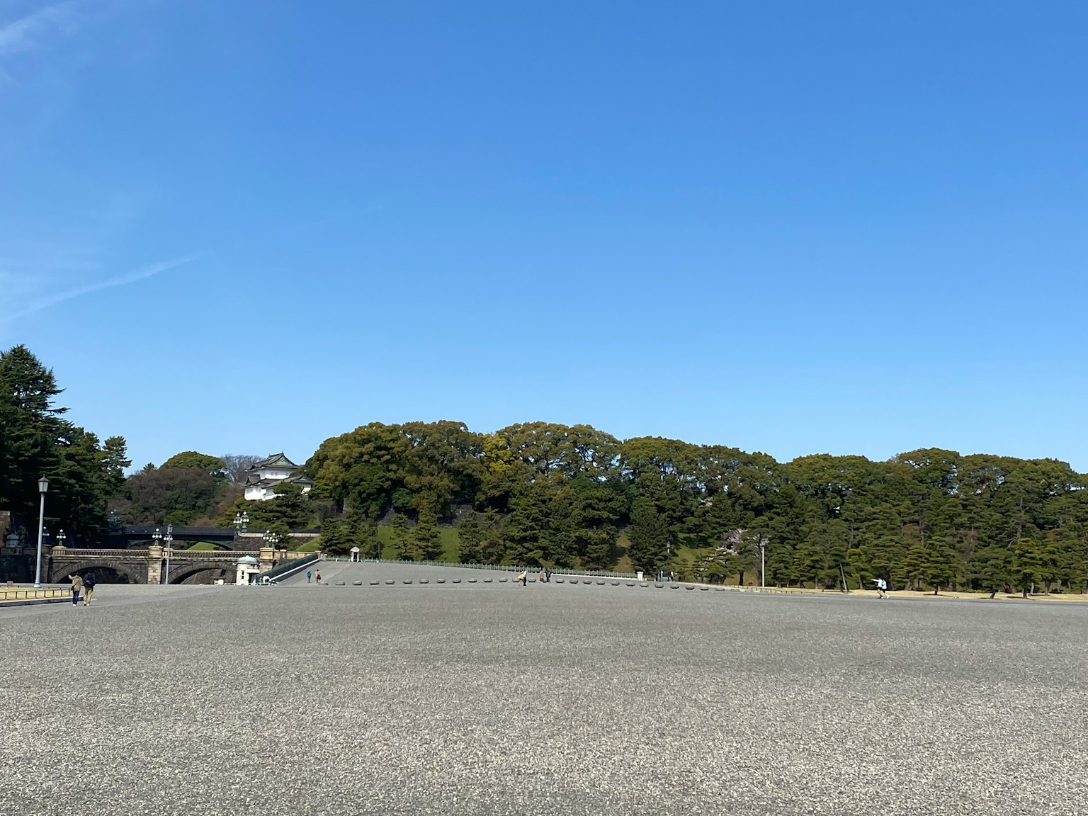 A wide gravel area under a clear blue sky with lush green trees in the background