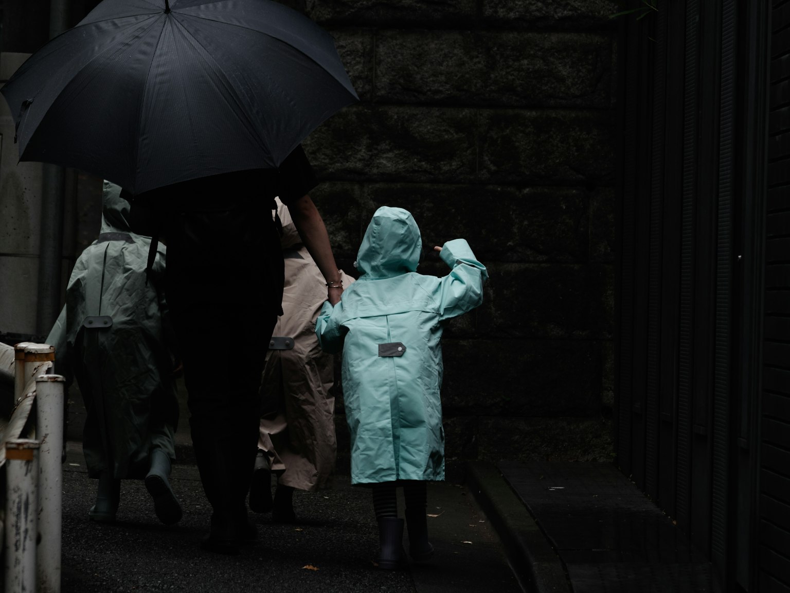 Familie, die unter einem schwarzen Regenschirm geht Kind im blauen Regenmantel und Erwachsene in hellen Regenmänteln