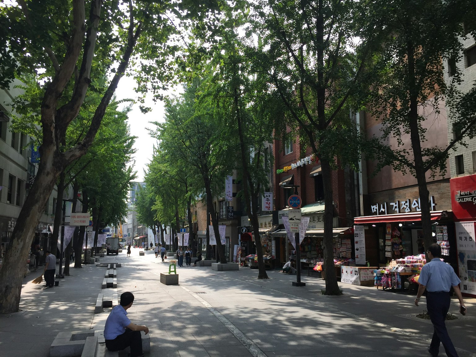 A vibrant street scene lined with lush trees featuring shops and people walking