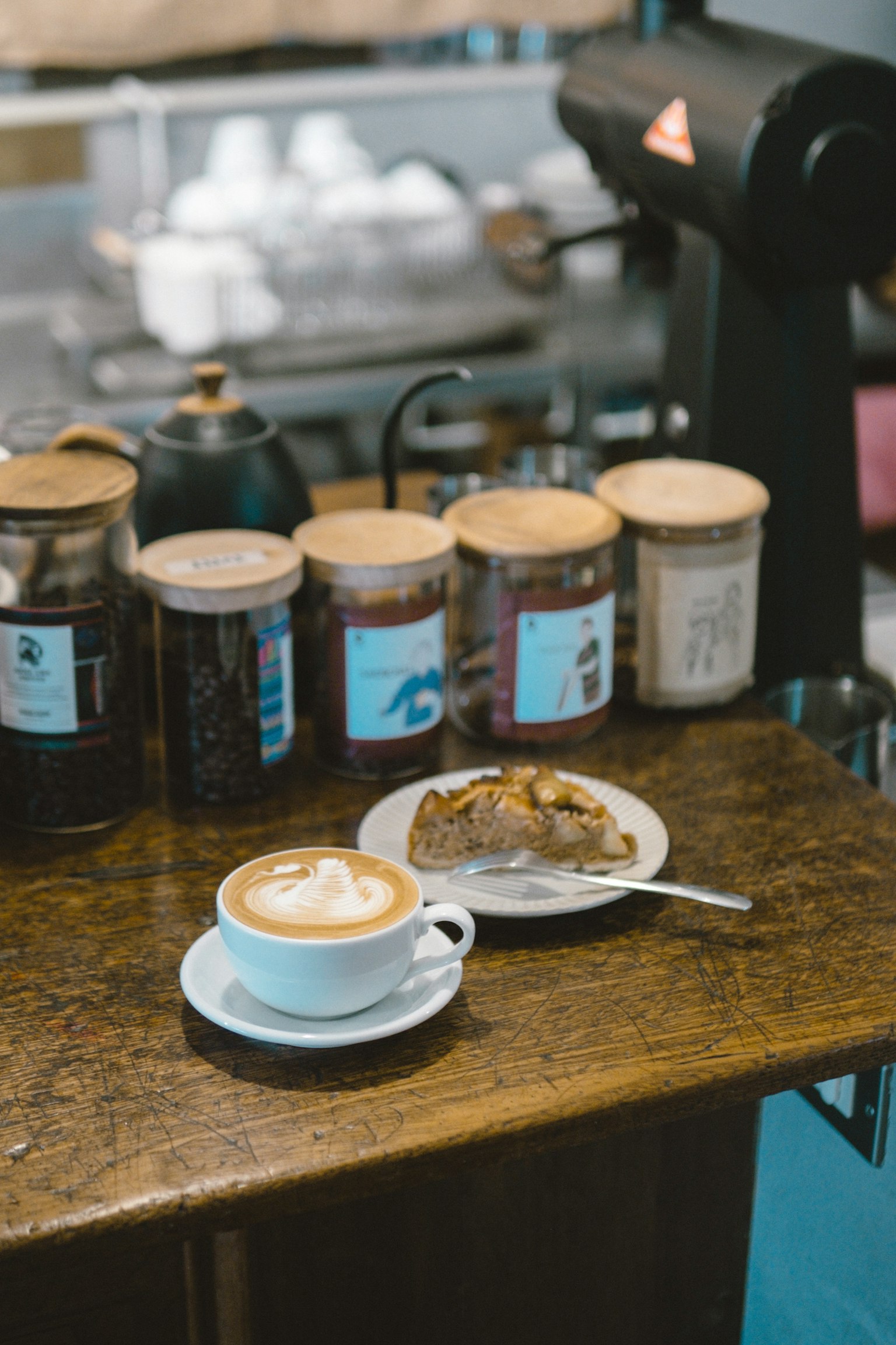 Una tazza di caffè con arte del latte e una fetta di torta su un piatto su un tavolo di legno con barattoli di chicchi di caffè sullo sfondo