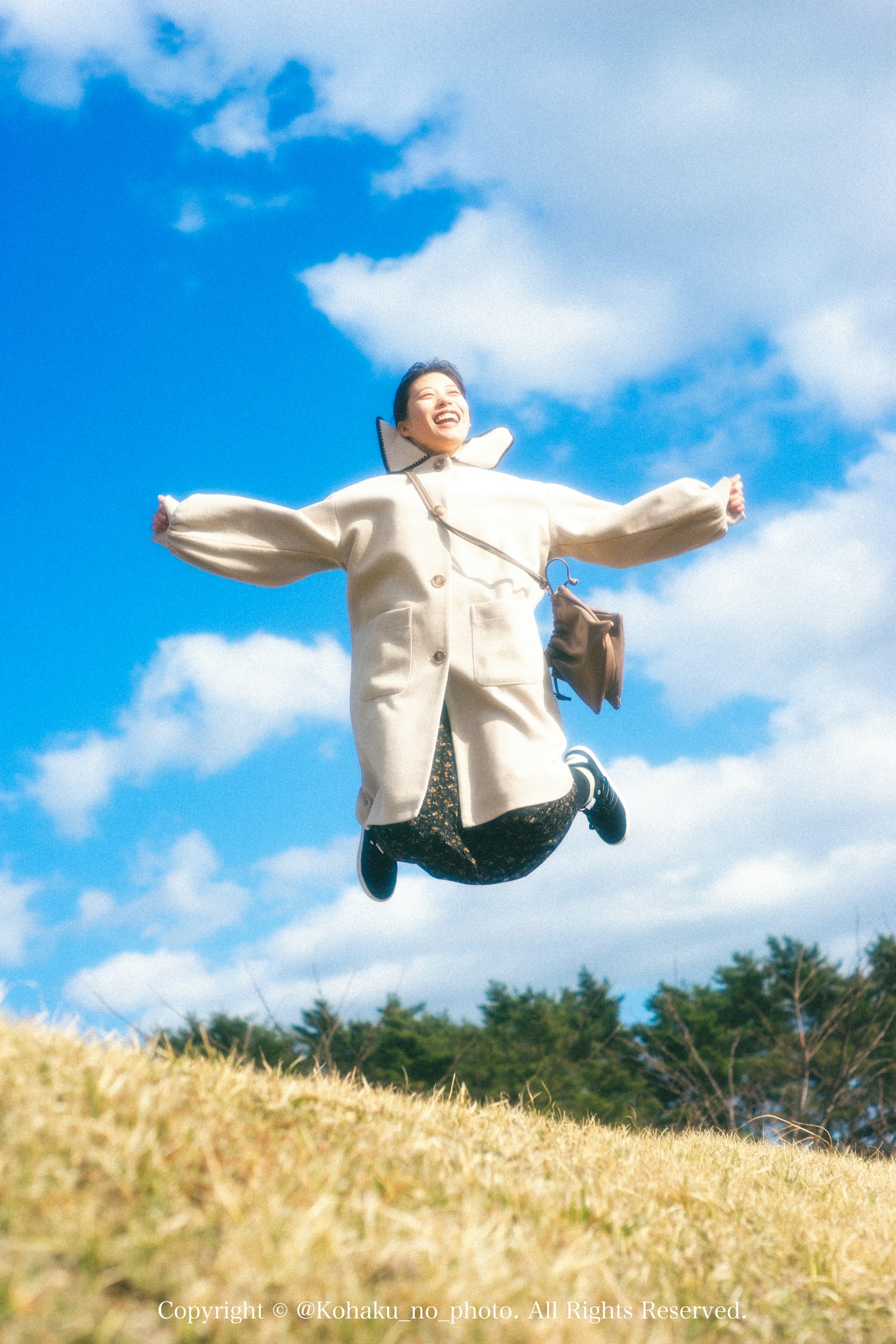 Woman jumping under a blue sky wearing a white coat and carrying a bag