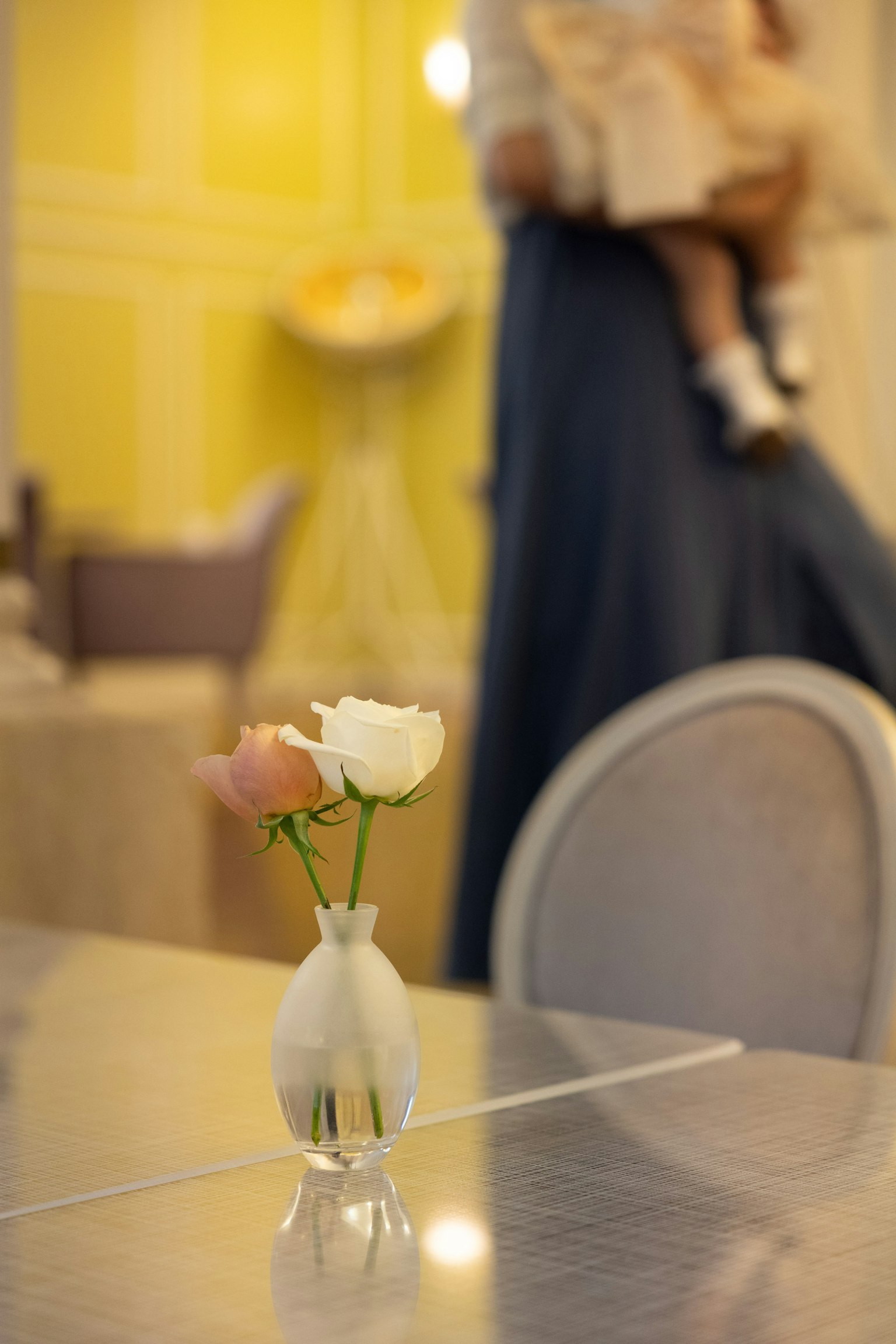 Eine kleine Vase auf einem Tisch mit rosa und weißen Blumen Eine Person, die ein Kind hält, ist im Hintergrund verschwommen